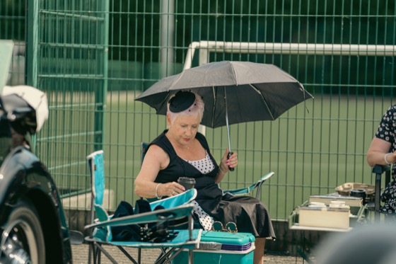 A woman in a black and white polka-dot dress sitting under a black umbrella near vintage vehicles.