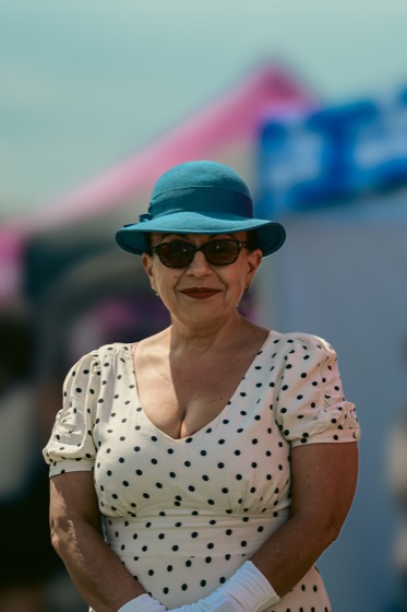A woman in a blue hat, sunglasses and polka-dot dress posing at a 1940s themed event.