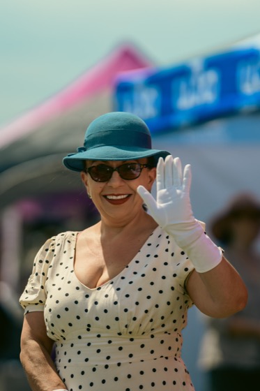 A woman in a blue hat and polka-dot dress smiling and waving at a 1940s themed event.