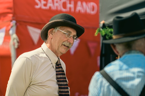 An older man in a bowler hat and round glasses talking at a 1940s themed event.
