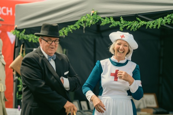 A costumed man in a dark suit and bowler hat laughing with a woman in a nurse-style costume.