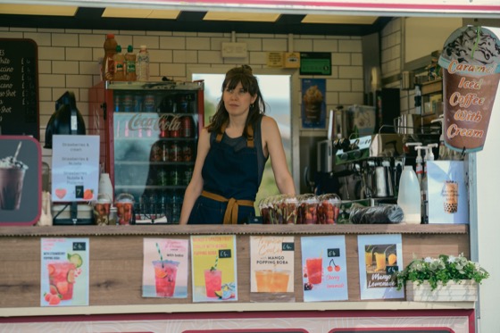 A drinks stall worker standing among signs, bottles and fruit at a public event.