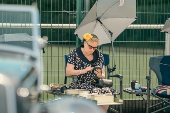 A woman in a floral dress sitting under a light umbrella and looking at a phone at a vintage-themed event.