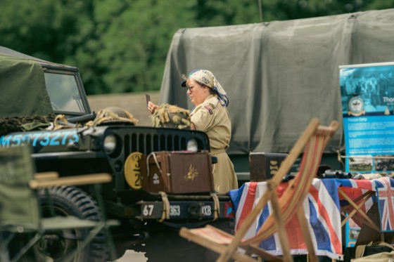 A woman in khaki period-style clothing using a phone beside a military jeep and Union Jack-covered table.