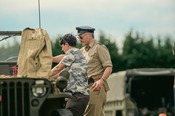 A man in khaki military-style uniform talking beside a woman near a green military vehicle.