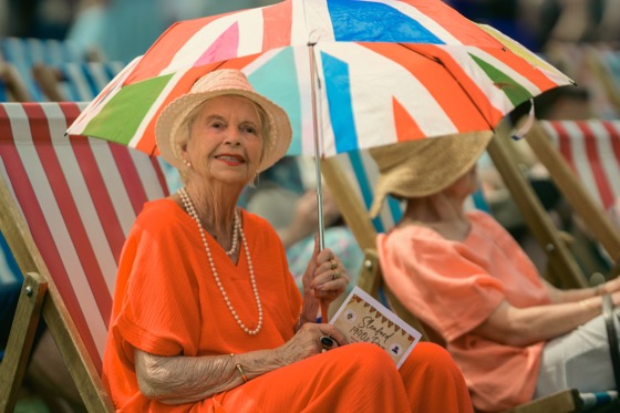 A woman in an orange outfit sitting under a Union Jack-coloured umbrella at an outdoor event.