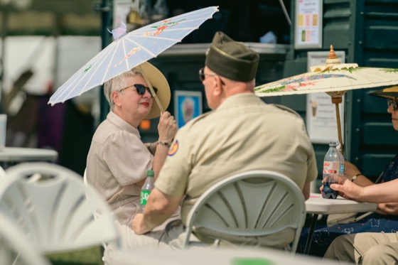 A woman with a white parasol talking with a man in military-style uniform at an outdoor table.