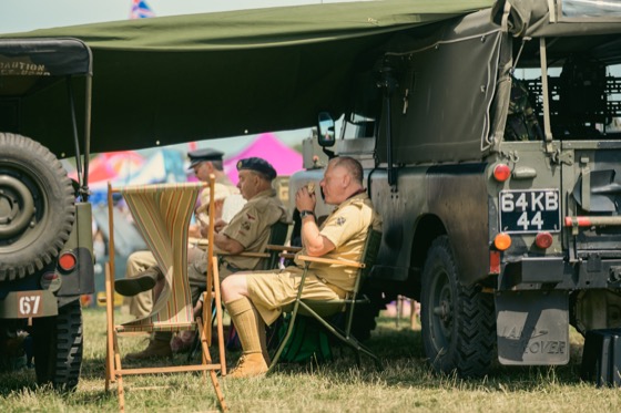 People in military-style clothing resting beside green military vehicles at a 1940s themed event.