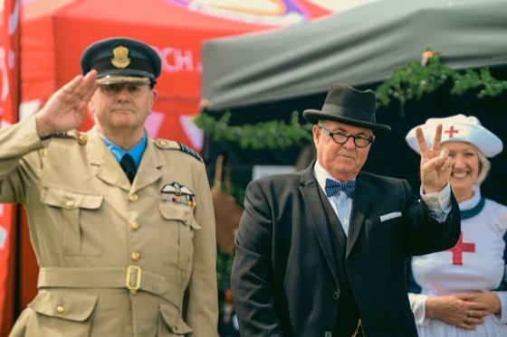 A man in RAF-style uniform saluting while another costumed man makes a V sign at a 1940s themed event.