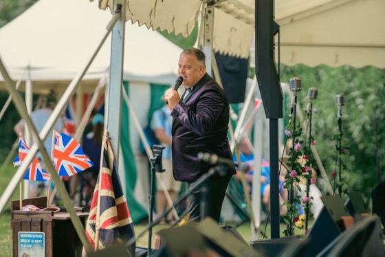 A male singer performing on stage with Union flags behind him at a 1940s themed event.
