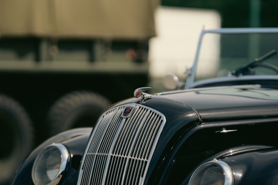 Vintage car bonnet badge and grille with a military vehicle blurred in the background.