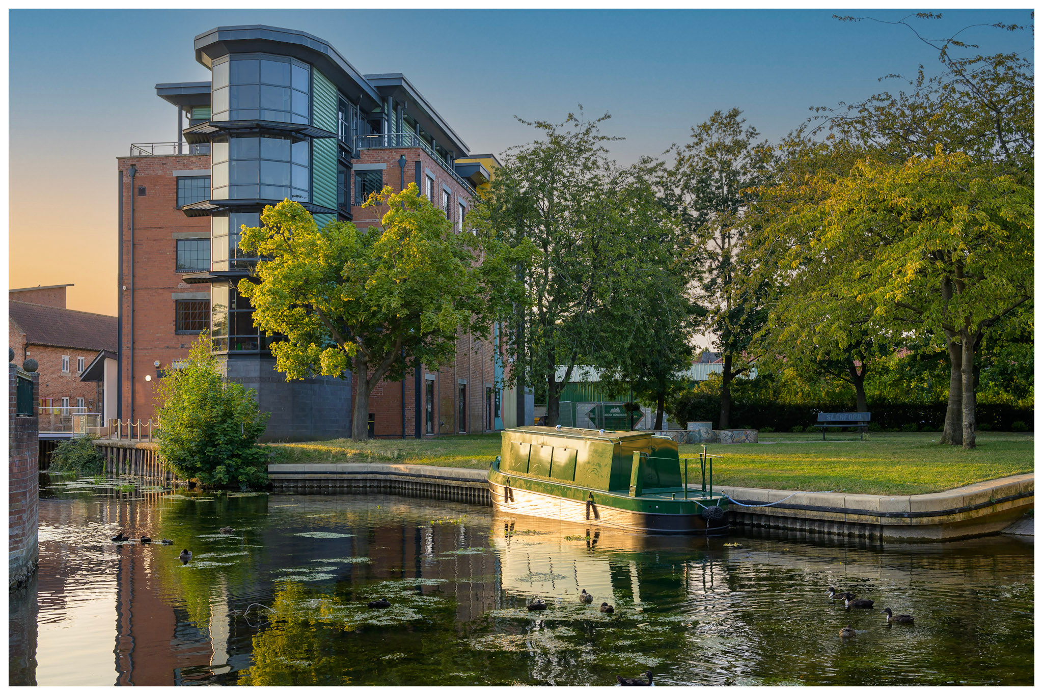 The River Slea at sunset beside the Hub, with a green narrowboat moored beneath trees and ducks on the water.