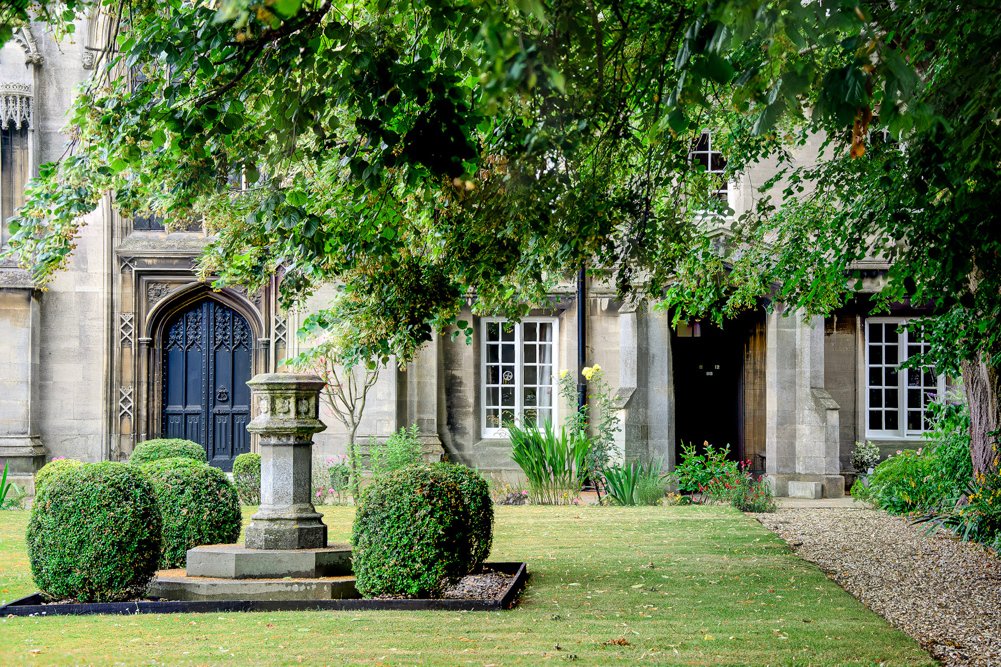 Carre's Hospital almshouses are seen through leafy trees, with arched doors, mullioned windows, clipped shrubs and a stone sundial in the garden.