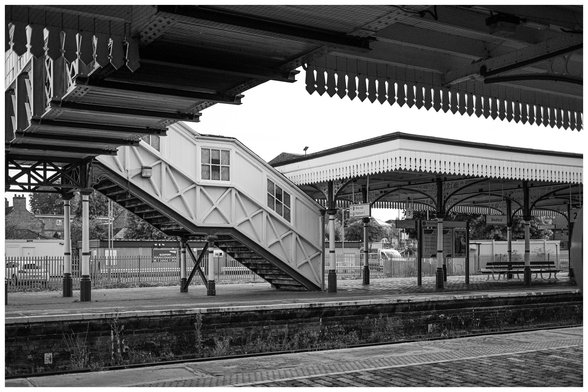 A black-and-white view across Sleaford railway station shows the enclosed footbridge, staircases, ornate canopy brackets and empty platforms.